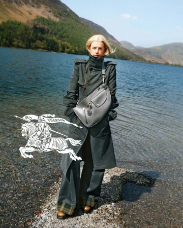 Blonde woman in dark coat standing by a lake wearing a crossbody bag for Burberry Winter 2024 campaign