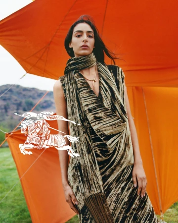 Model wearing a patterned olive scarf and dress standing in front of a bright orange tent structure outdoors.