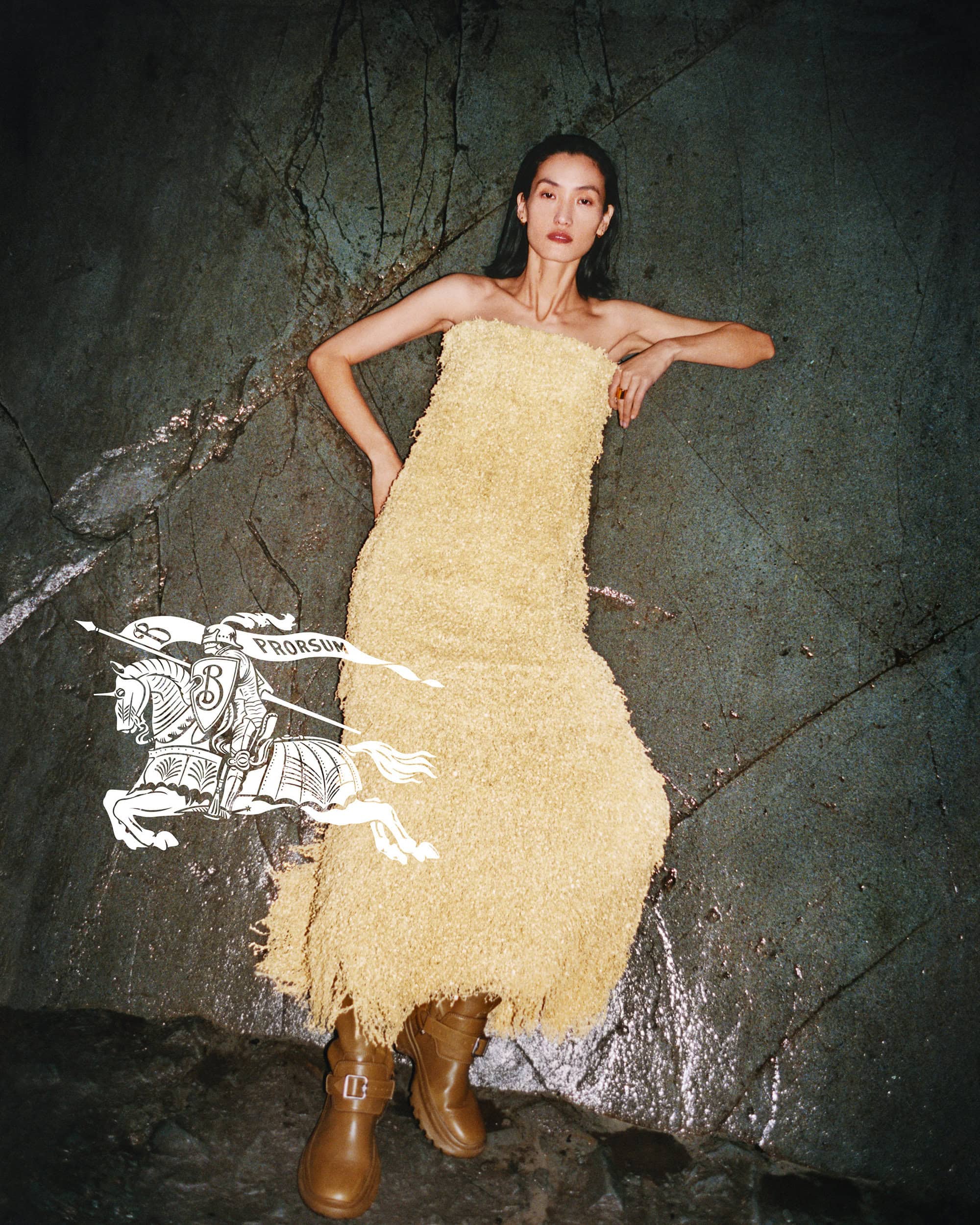 Model Lina Zhang in a textured yellow strapless dress and brown boots leaning against a rocky stone wall for Burberry Winter 2024 campaign