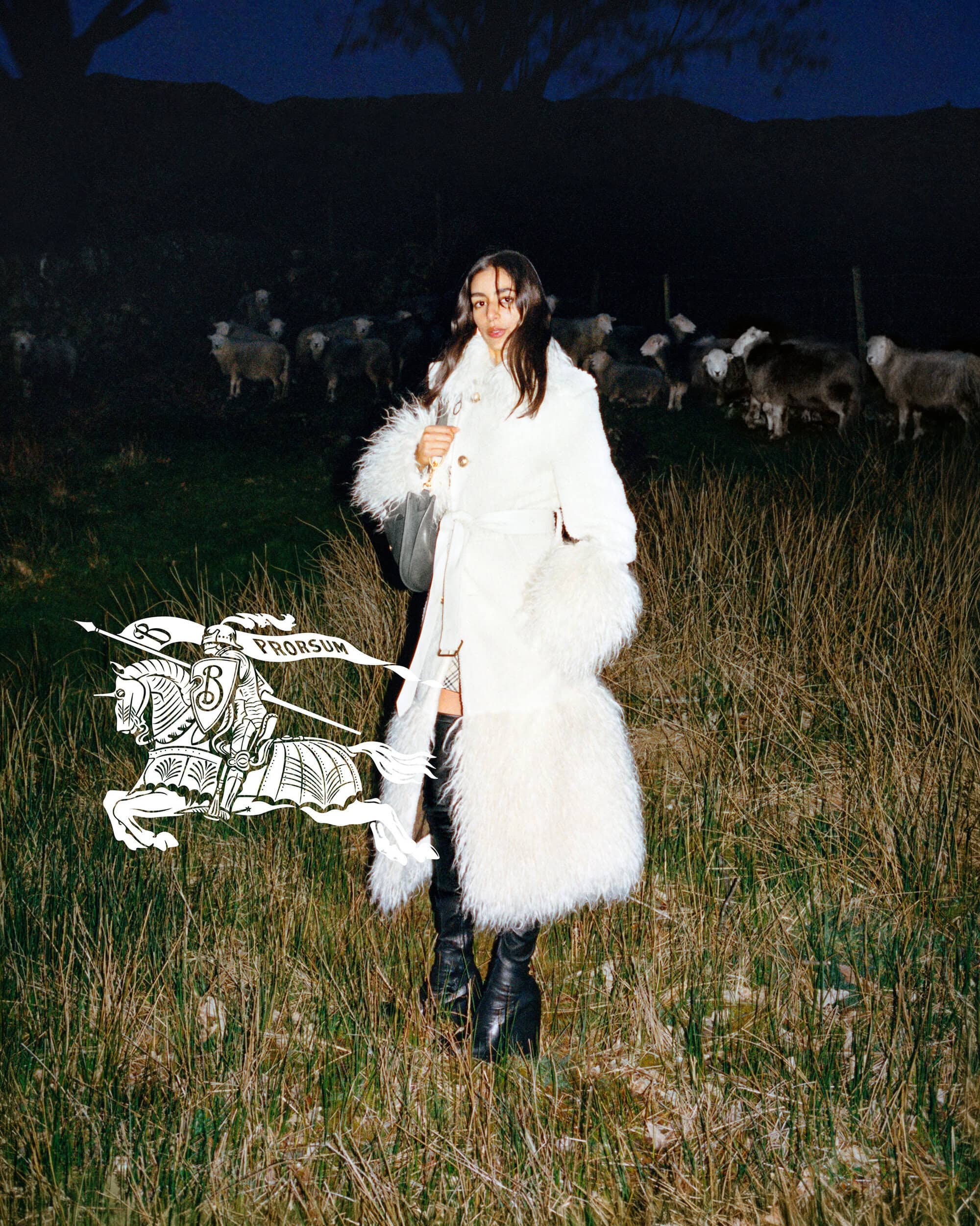 Model in a white belted coat with fur trim standing in a field at night with sheep grazing in the background.