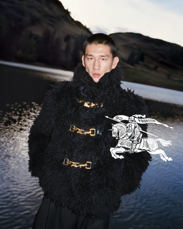 East Asian male model wearing a black textured fleece jacket with gold hardware standing before a lake and mountain backdrop