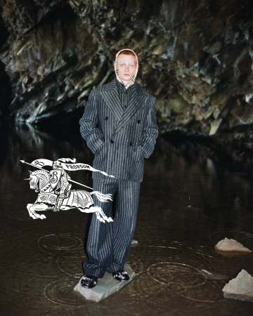 Model standing on a rock in shallow water wearing a pinstripe suit and hooded scarf, with a large overlaid Burberry knight logo.