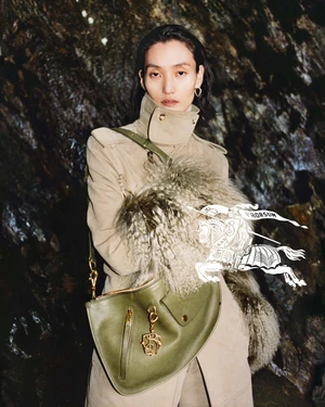 Model wearing a beige coat and holding an olive green handbag against a rocky background
