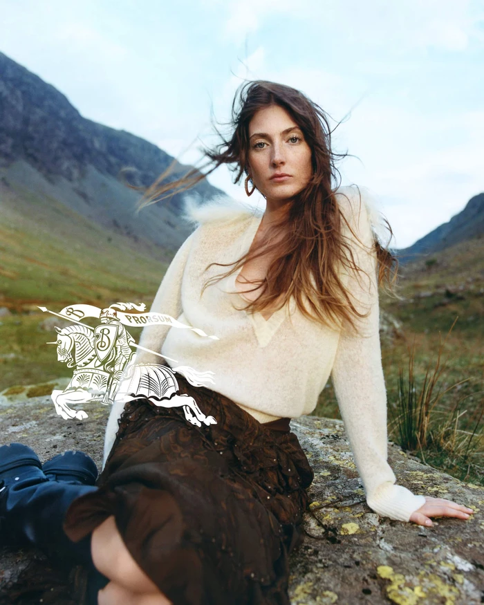 Model in cream knit sweater and dark skirt seated outdoors against a mountainous landscape for Burberry