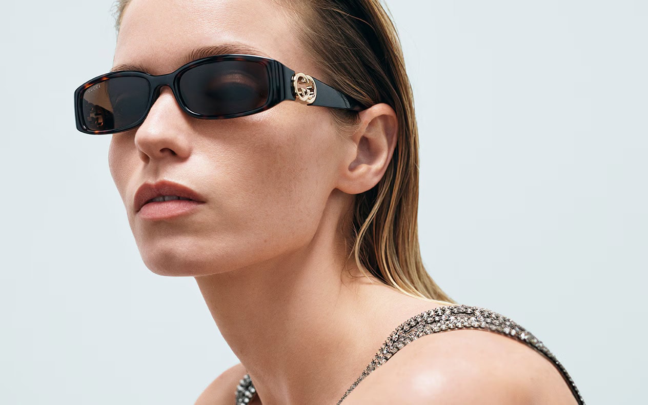 Model wearing dark rectangular sunglasses with gold hardware against a light background for Gucci