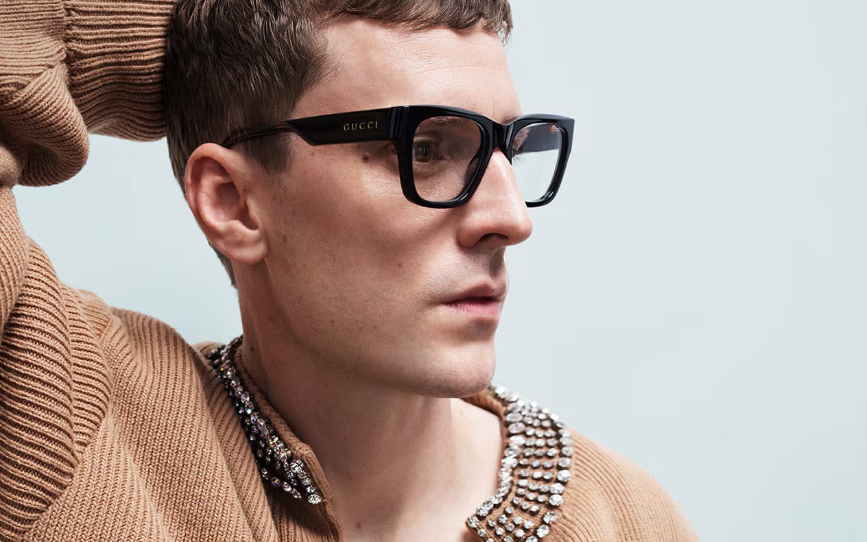 Man wearing black Gucci optical frames and a textured brown sweater against a neutral background for the Eyewear Fall 2024 campaign.