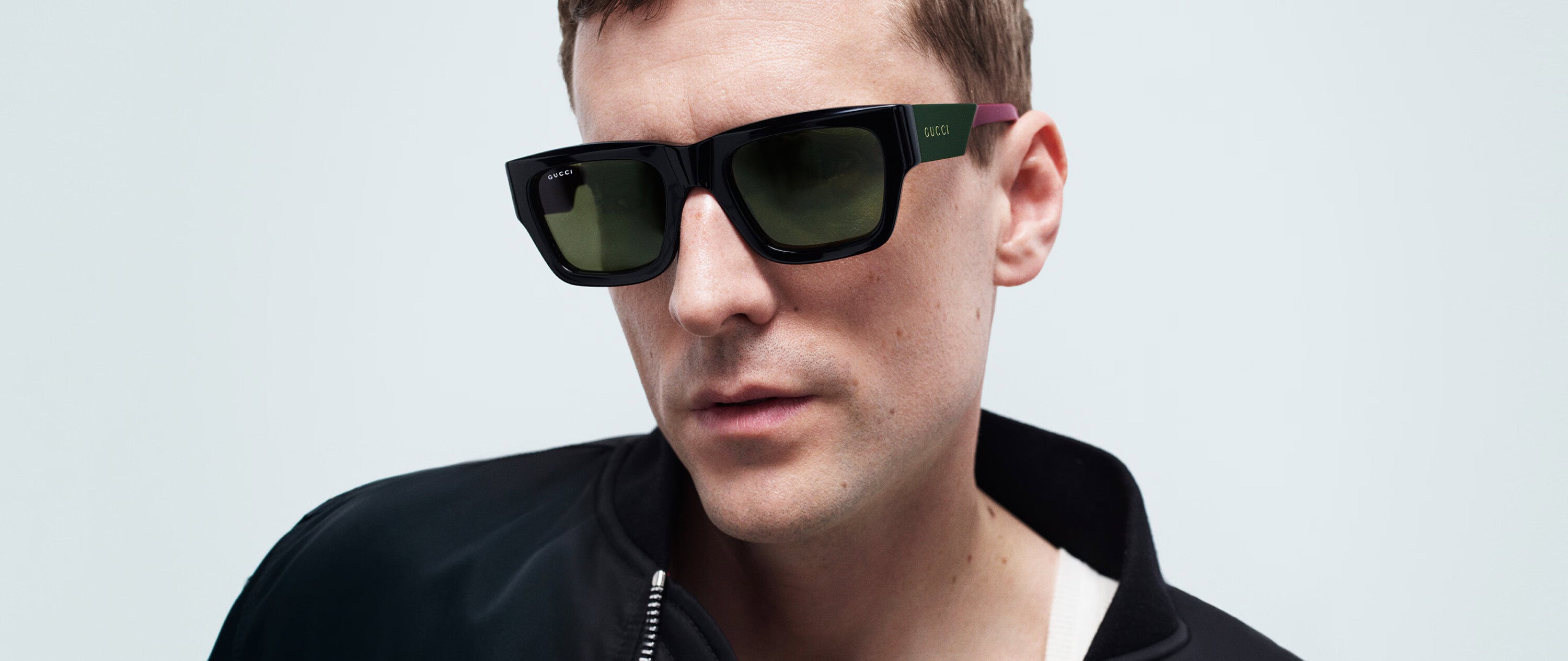 Close-up portrait of a man wearing black Gucci sunglasses and a black zip-up jacket against a plain white background.