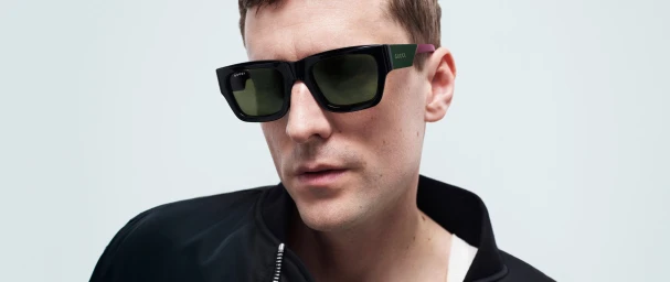 Close-up portrait of a man wearing black Gucci sunglasses and a black zip-up jacket against a plain white background.