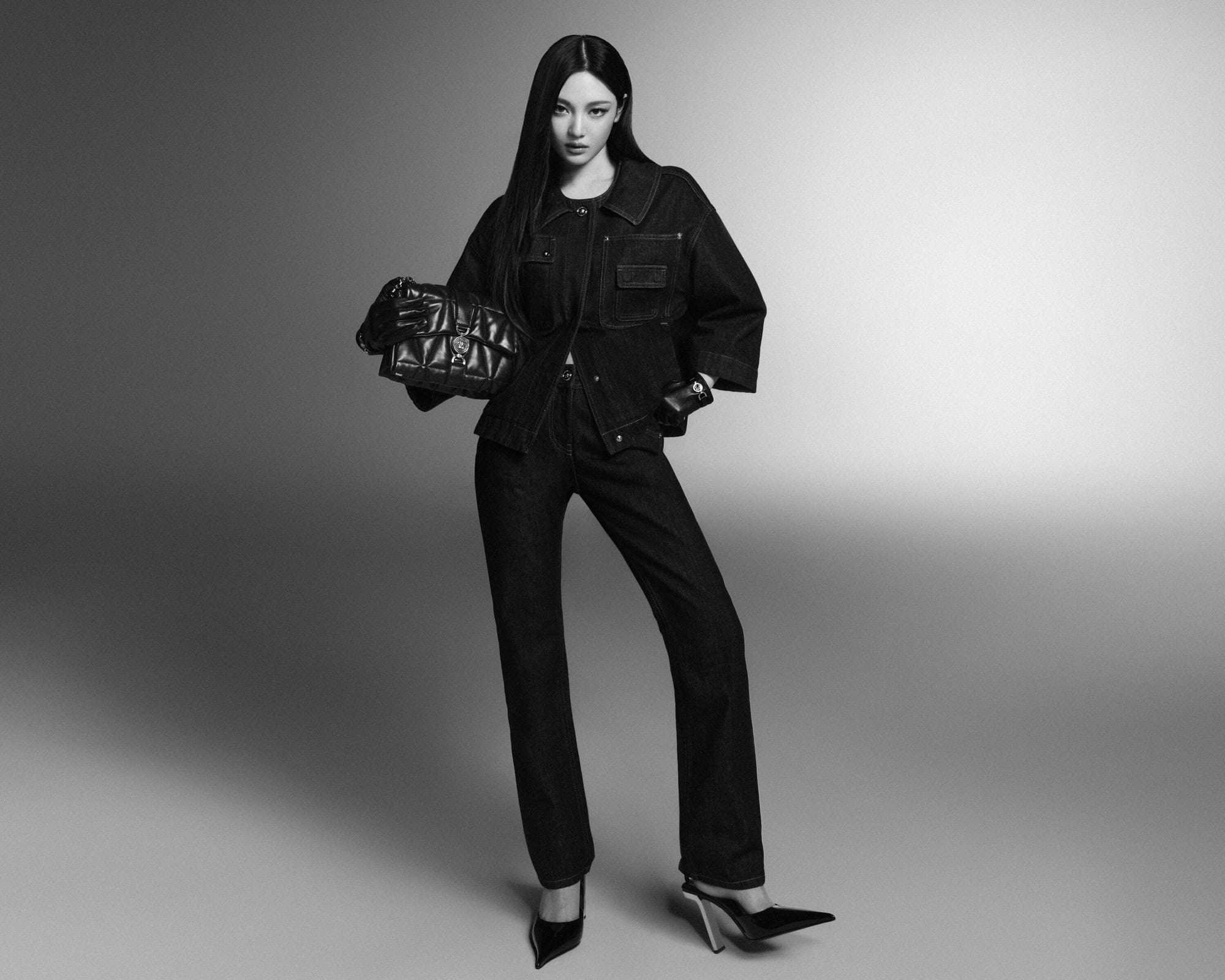 Model standing against a seamless background holding a quilted leather handbag and wearing a dark denim jacket and matching trousers.