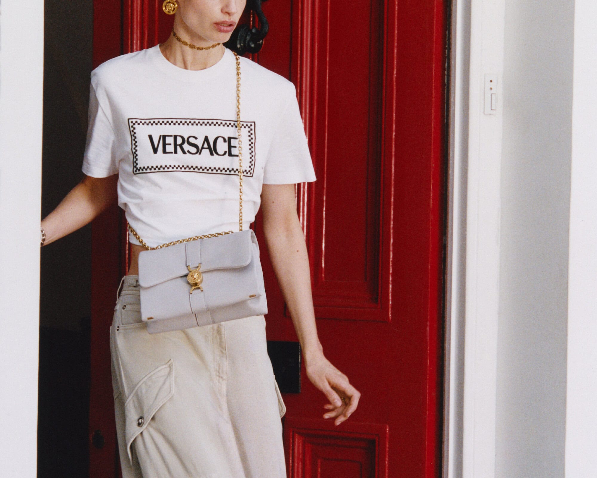 Model wearing a white Versace logo t-shirt and light beige cargo pants with a grey crossbody bag in front of a red door