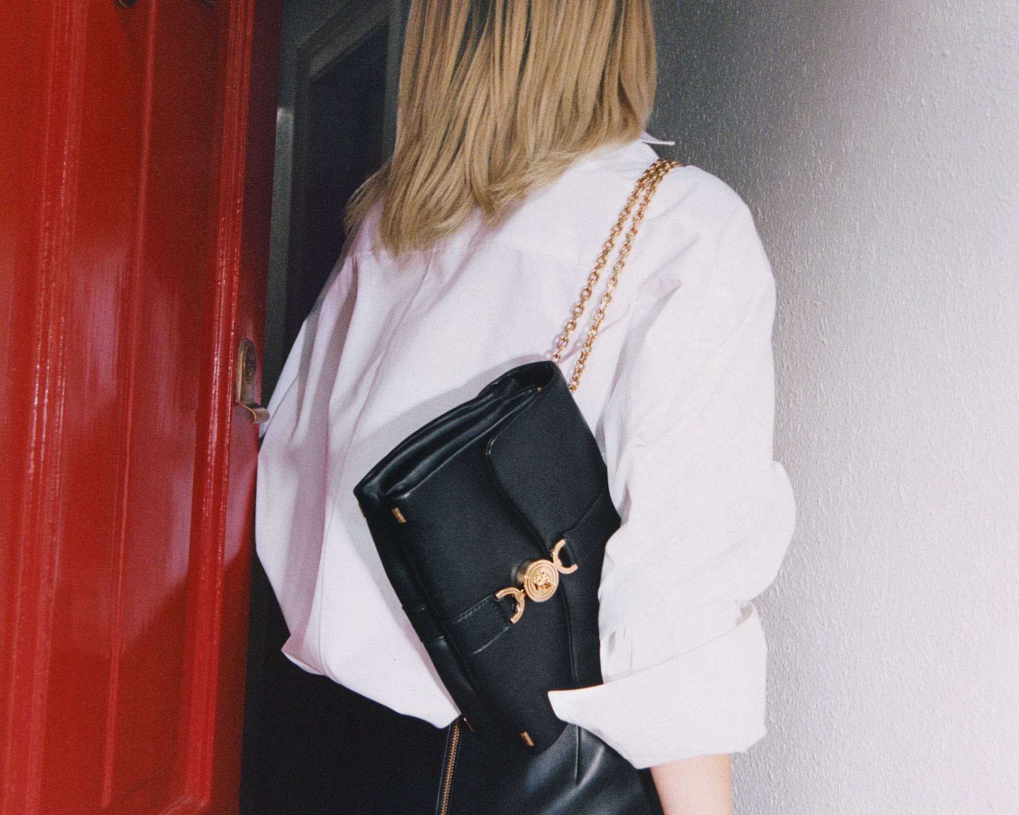 Model with blonde hair wearing a white button-down shirt and carrying a black leather shoulder bag with gold hardware.