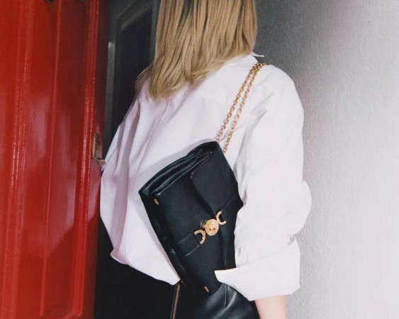 Model with blonde hair wearing a white button-down shirt and carrying a black leather shoulder bag with gold hardware.
