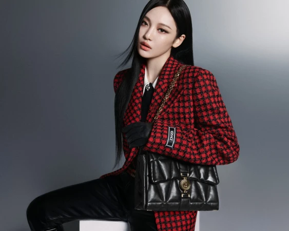 Model with long dark hair wearing a red plaid tweed blazer and holding a black quilted leather handbag against a grey backdrop