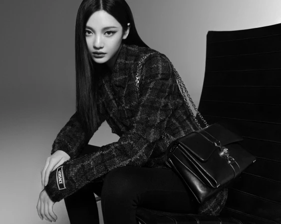 Model Ningning wearing a textured jacket with a black handbag seated against a grey studio backdrop