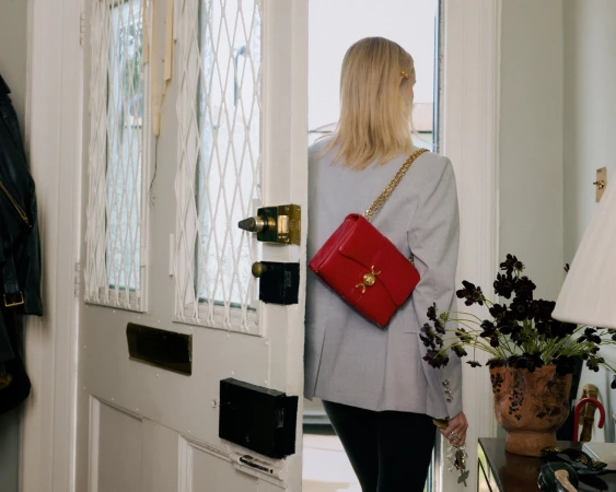 Blonde woman wearing a grey blazer and carrying a bright red shoulder bag while opening a white front door.