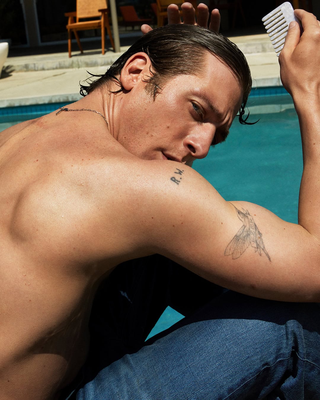 Shirtless man with wet slicked hair holding a white comb beside a blue outdoor swimming pool