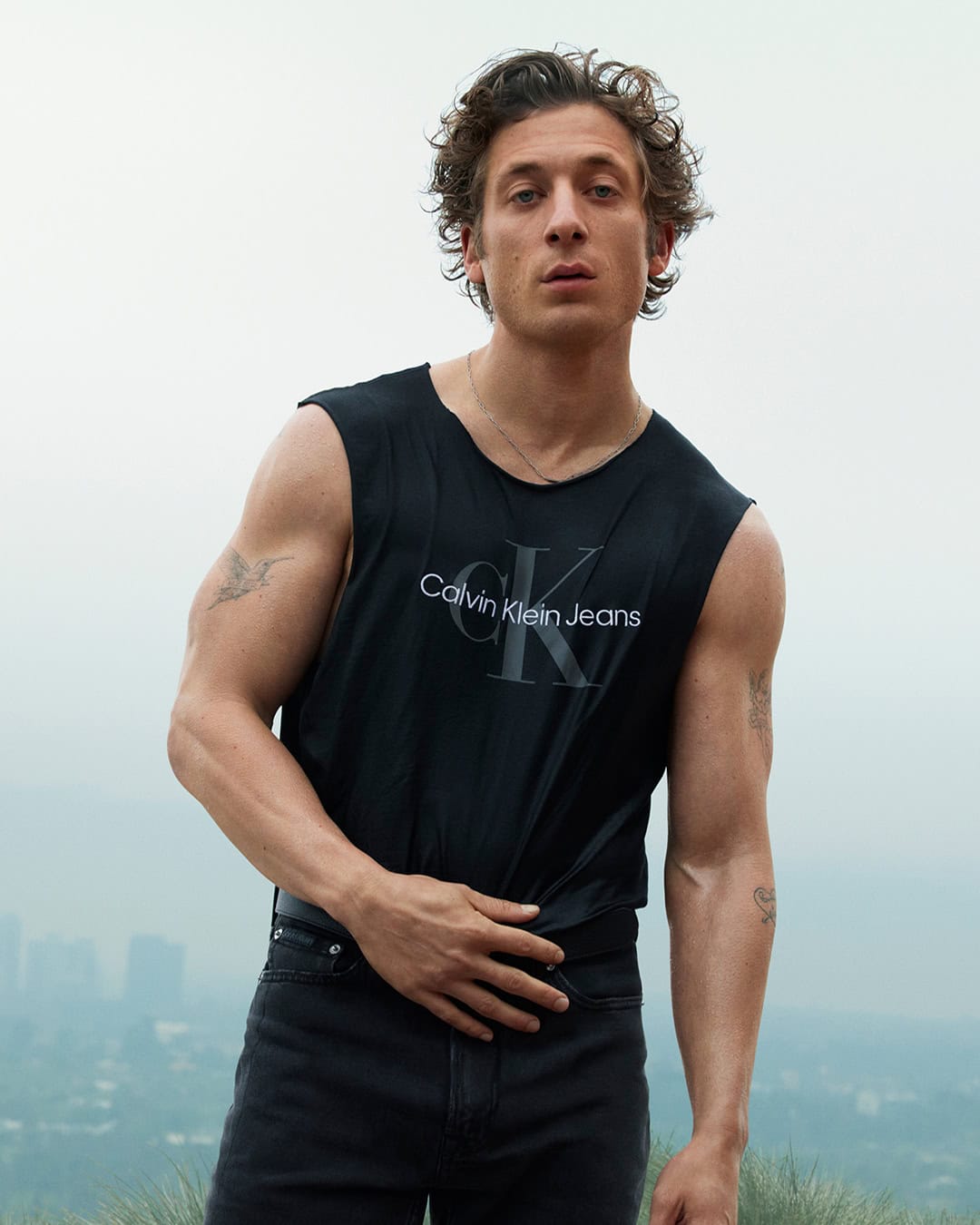 Jeremy Allen White wearing a black Calvin Klein sleeveless tank top against an overcast sky and distant cityscape.