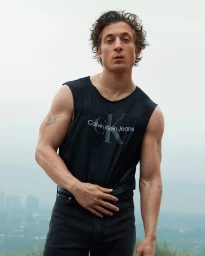 Jeremy Allen White wearing a black Calvin Klein sleeveless tank top against an overcast sky and distant cityscape.