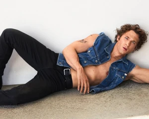Jeremy Allen White lying down wearing a denim vest and black jeans against a neutral wall
