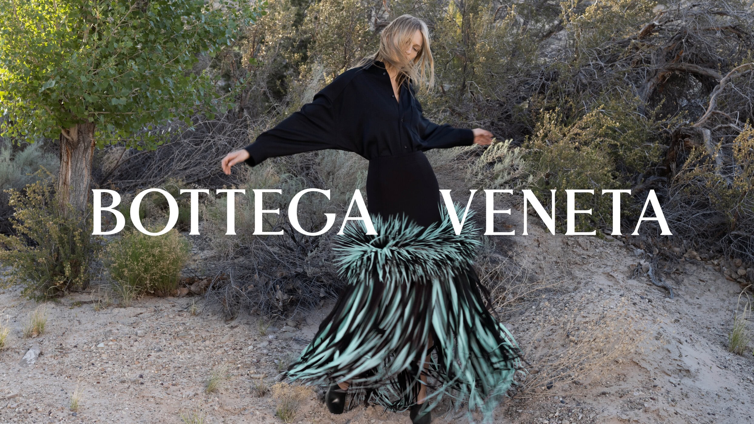 Model in black button-down shirt and fringed skirt posing outdoors in a sparse landscape with trees and brush.