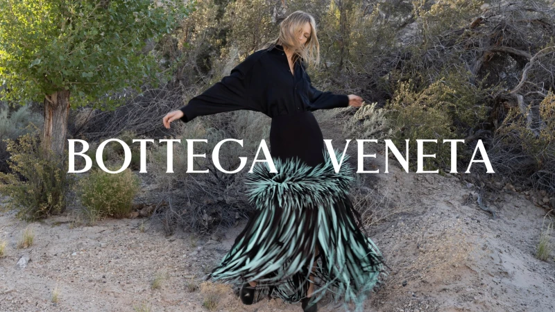 Model in black button-down shirt and fringed skirt posing outdoors in a sparse landscape with trees and brush.