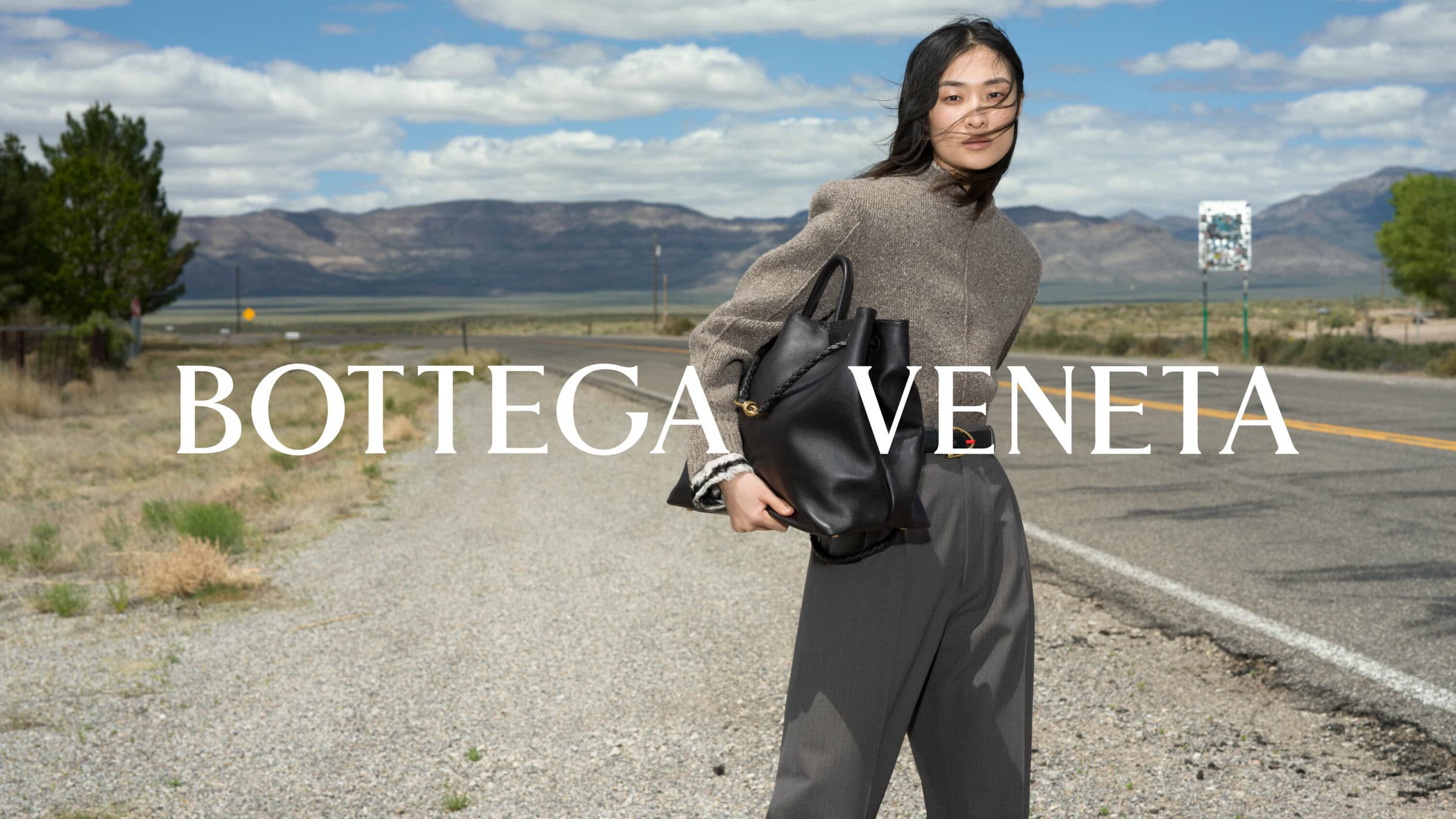 Model standing on a roadside holding a large black leather handbag wearing a textured grey top
