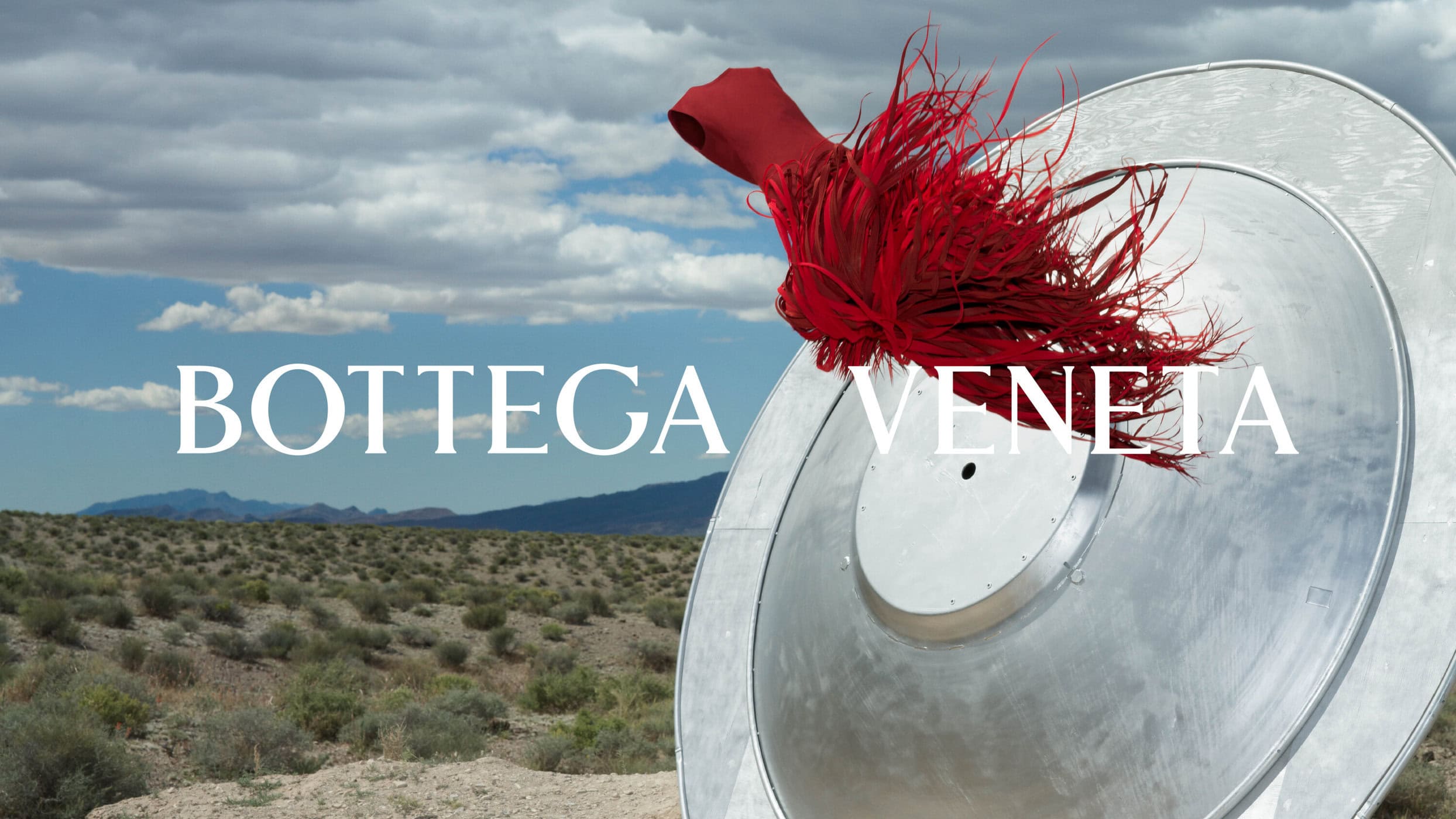 Vibrant red fringed garment emerging from a large metallic disc in an expansive desert landscape under a partly cloudy sky.