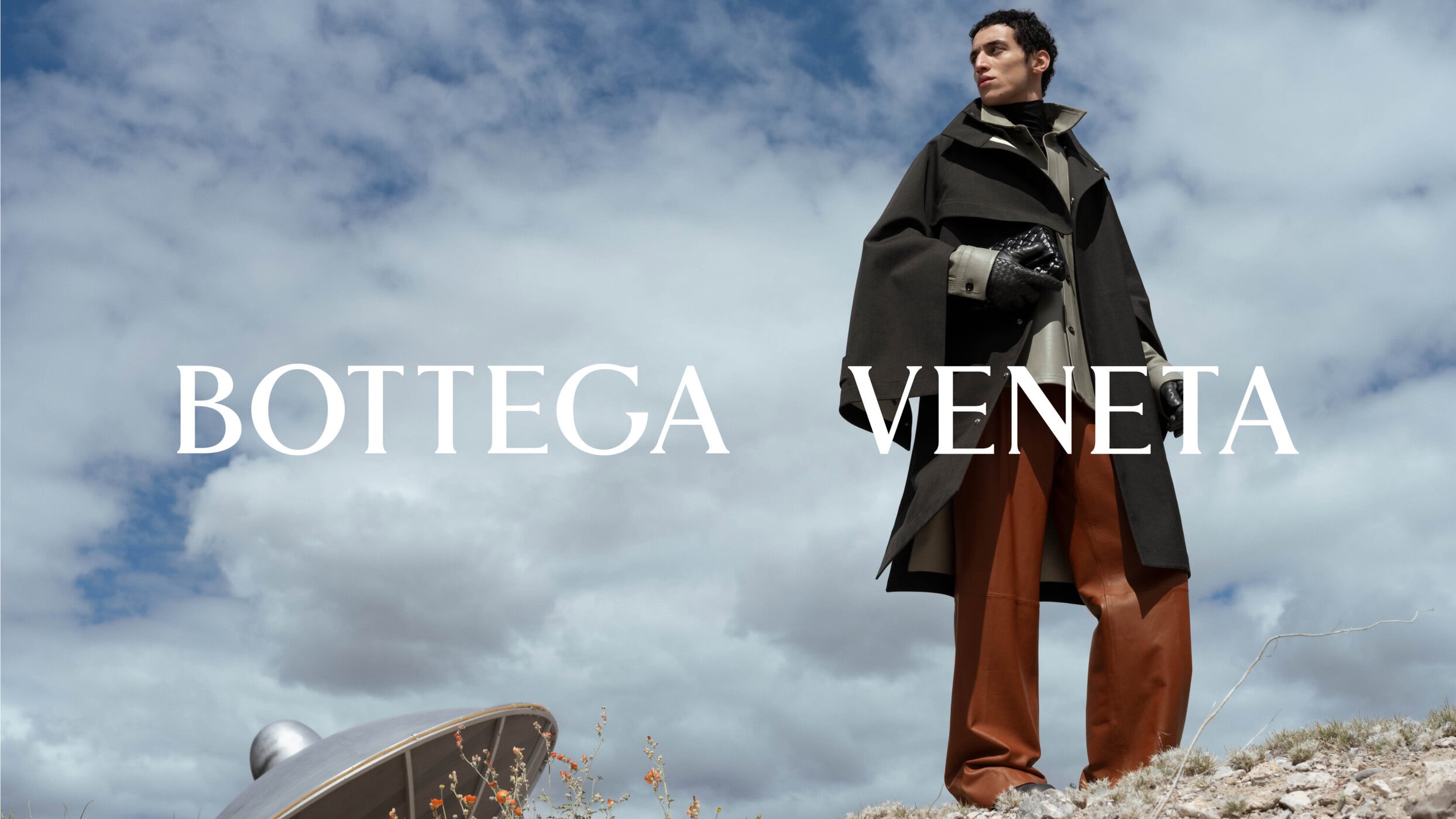 Man wearing a dark coat and brown trousers standing against an outdoor sky backdrop for Bottega Veneta