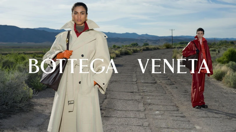 Model wearing a light beige belted trench coat standing on a dusty road with a second model in red apparel in the background