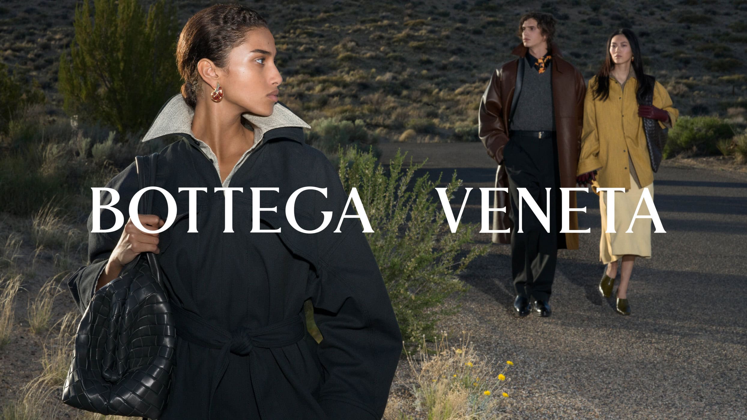 Woman in a black belted coat holding a woven leather handbag outdoors, with two figures walking on a desert road behind her.