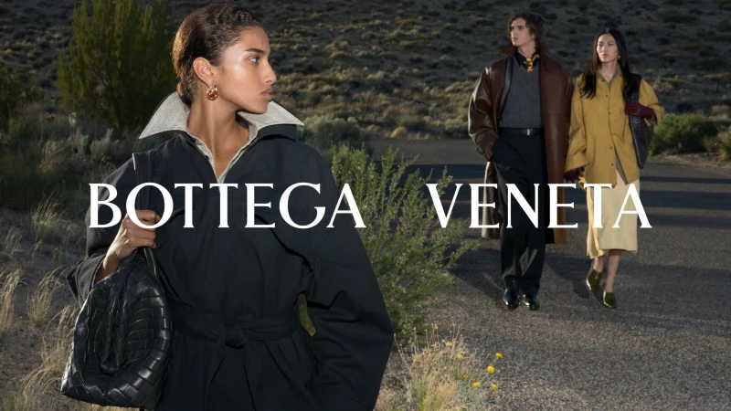 Woman in a black belted coat holding a woven leather handbag outdoors, with two figures walking on a desert road behind her.