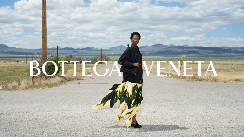 Model standing on a gravel road wearing a black shirt and patterned skirt with mountains and blue sky in the background