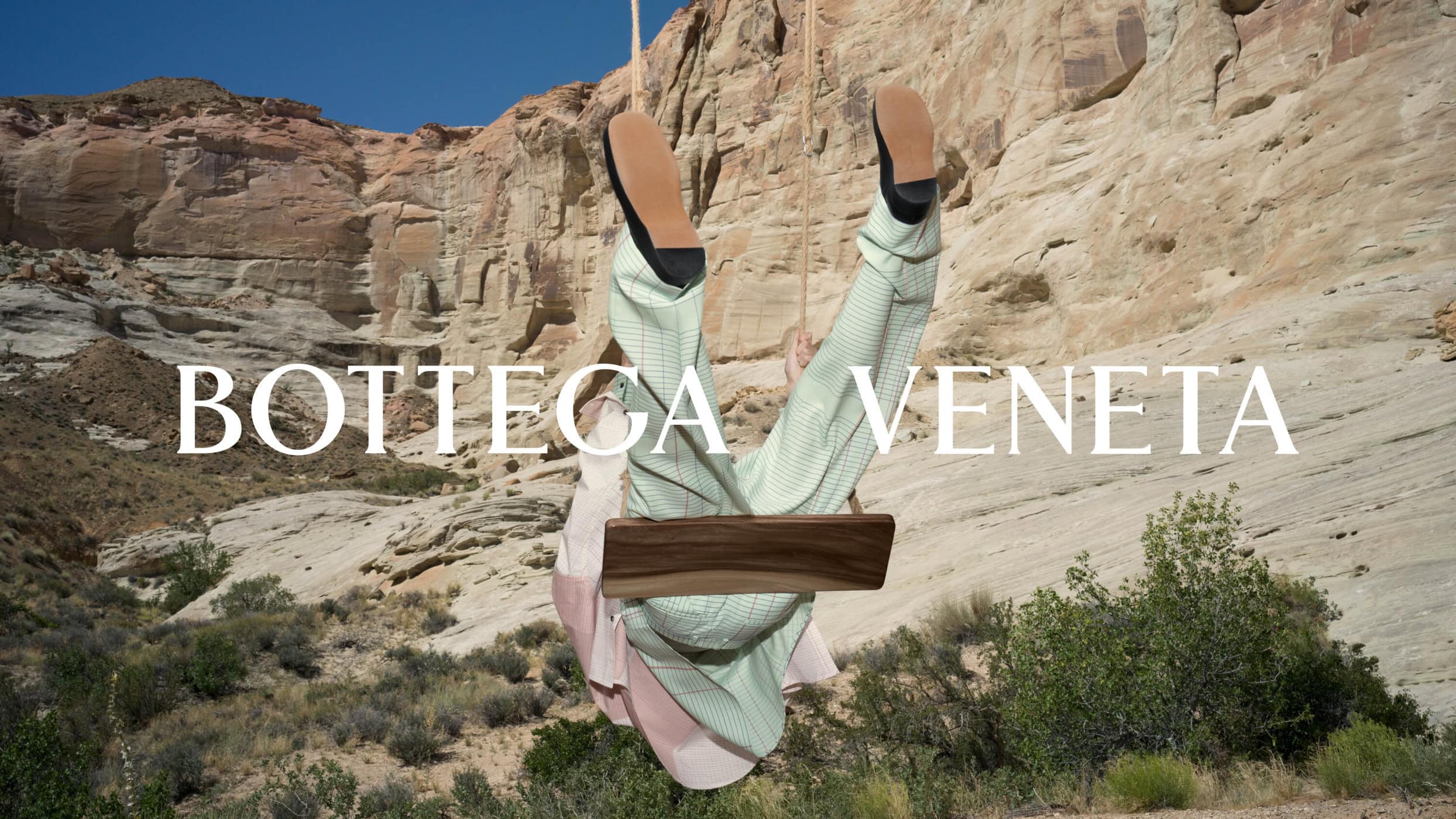 Person hanging upside down on a wooden swing against a rugged rocky desert canyon landscape with green shrubs.