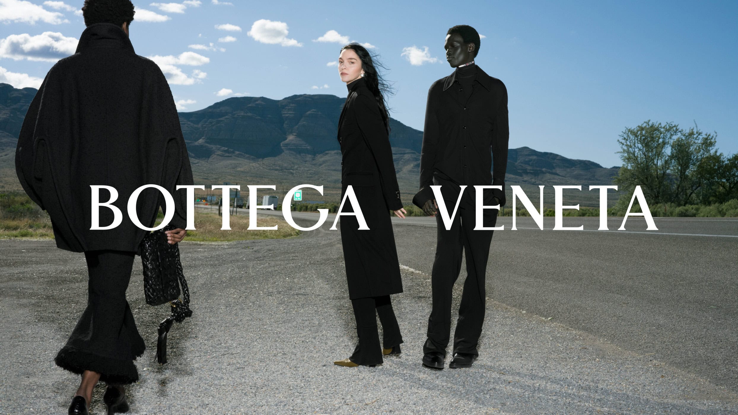 Three models wearing black clothing standing along an asphalt roadside with distant mountains and a cloudy blue sky.