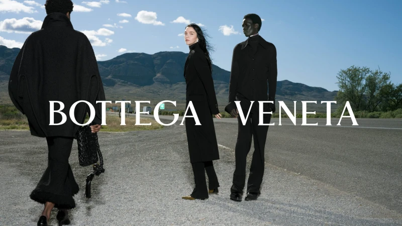 Three models wearing black clothing standing along an asphalt roadside with distant mountains and a cloudy blue sky.