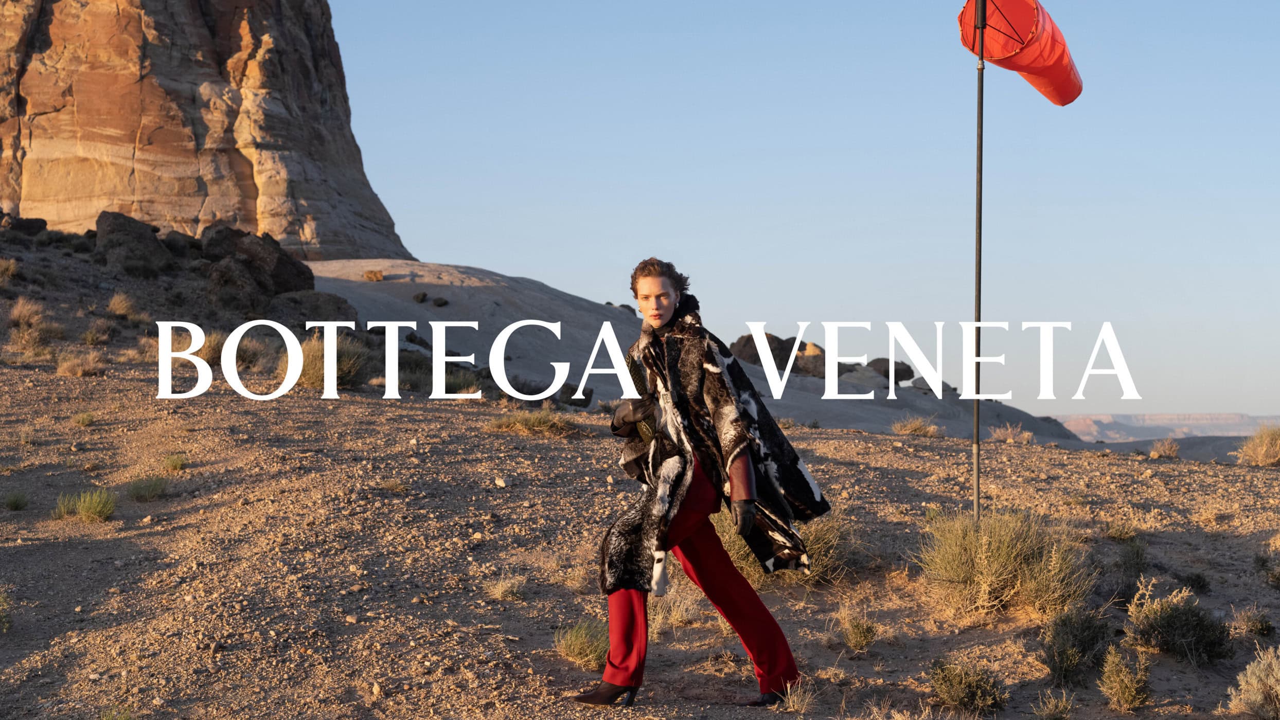 Model wearing a patterned coat and red trousers walks across a desert landscape near a red windsock under clear sky.