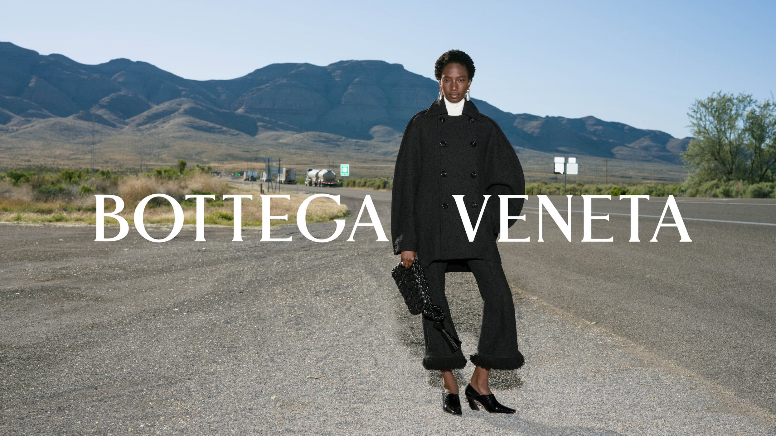 Model standing on asphalt road wearing black coat holding dark handbag with mountains in the background
