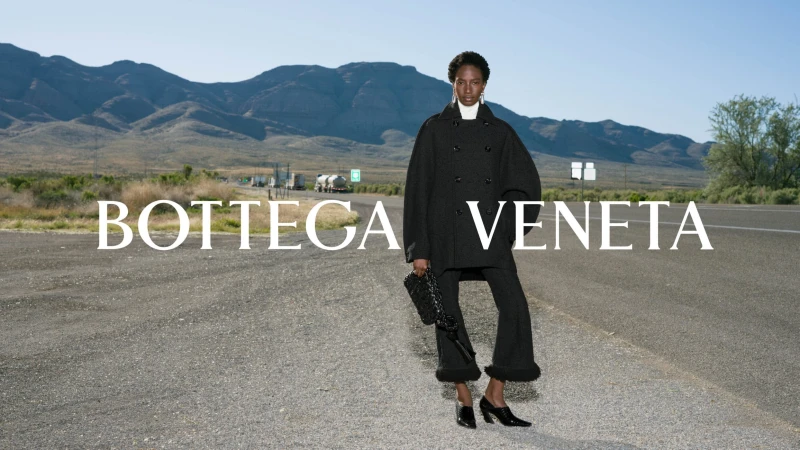 Model standing on asphalt road wearing black coat holding dark handbag with mountains in the background