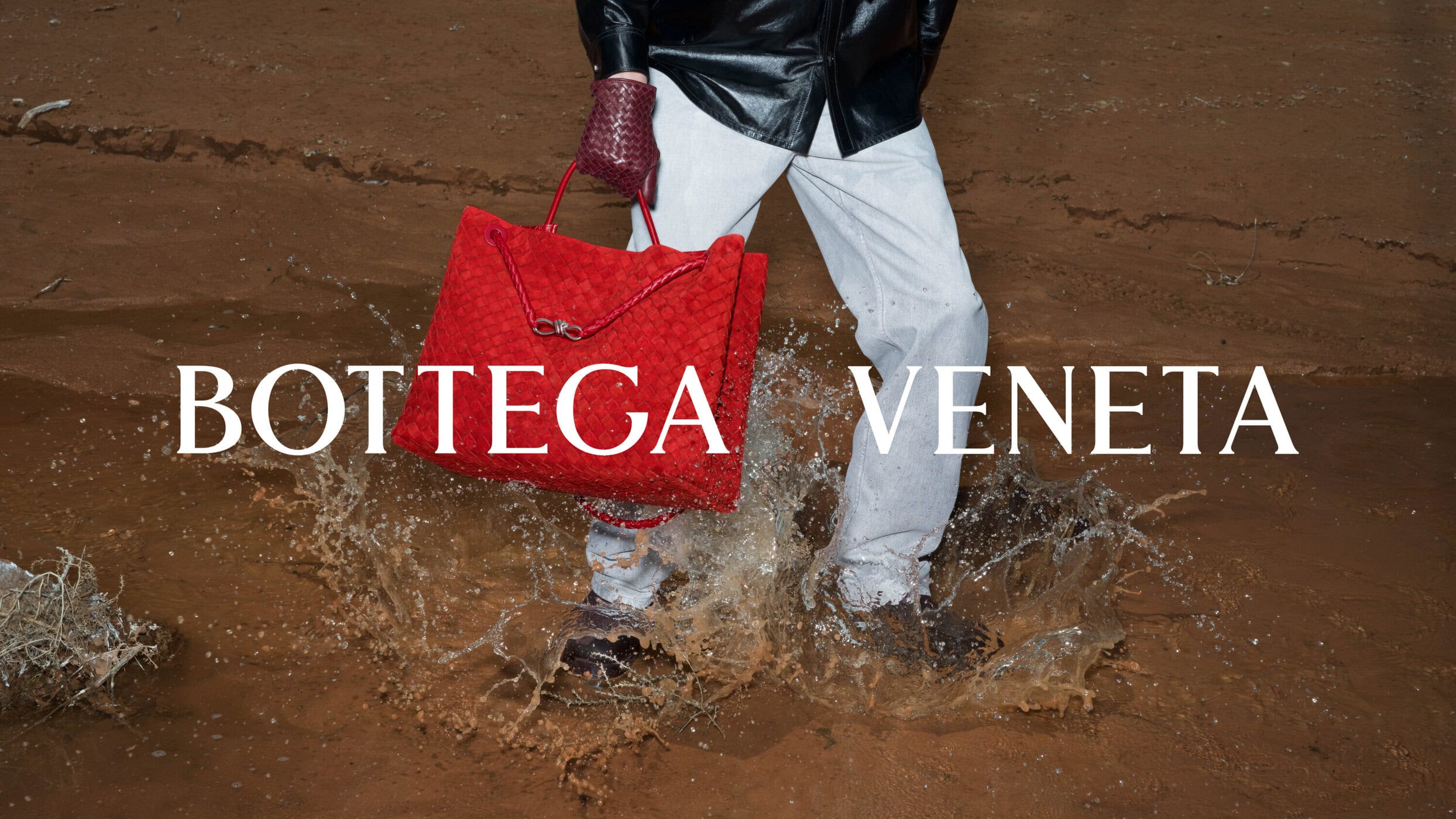 Model splashing through mud in light trousers and a black leather jacket carrying a large red woven leather bag for Bottega Veneta.
