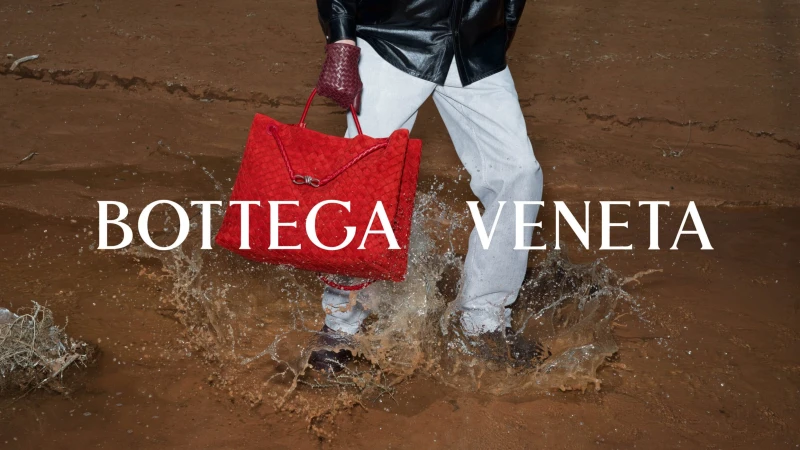 Model splashing through mud in light trousers and a black leather jacket carrying a large red woven leather bag for Bottega Veneta.