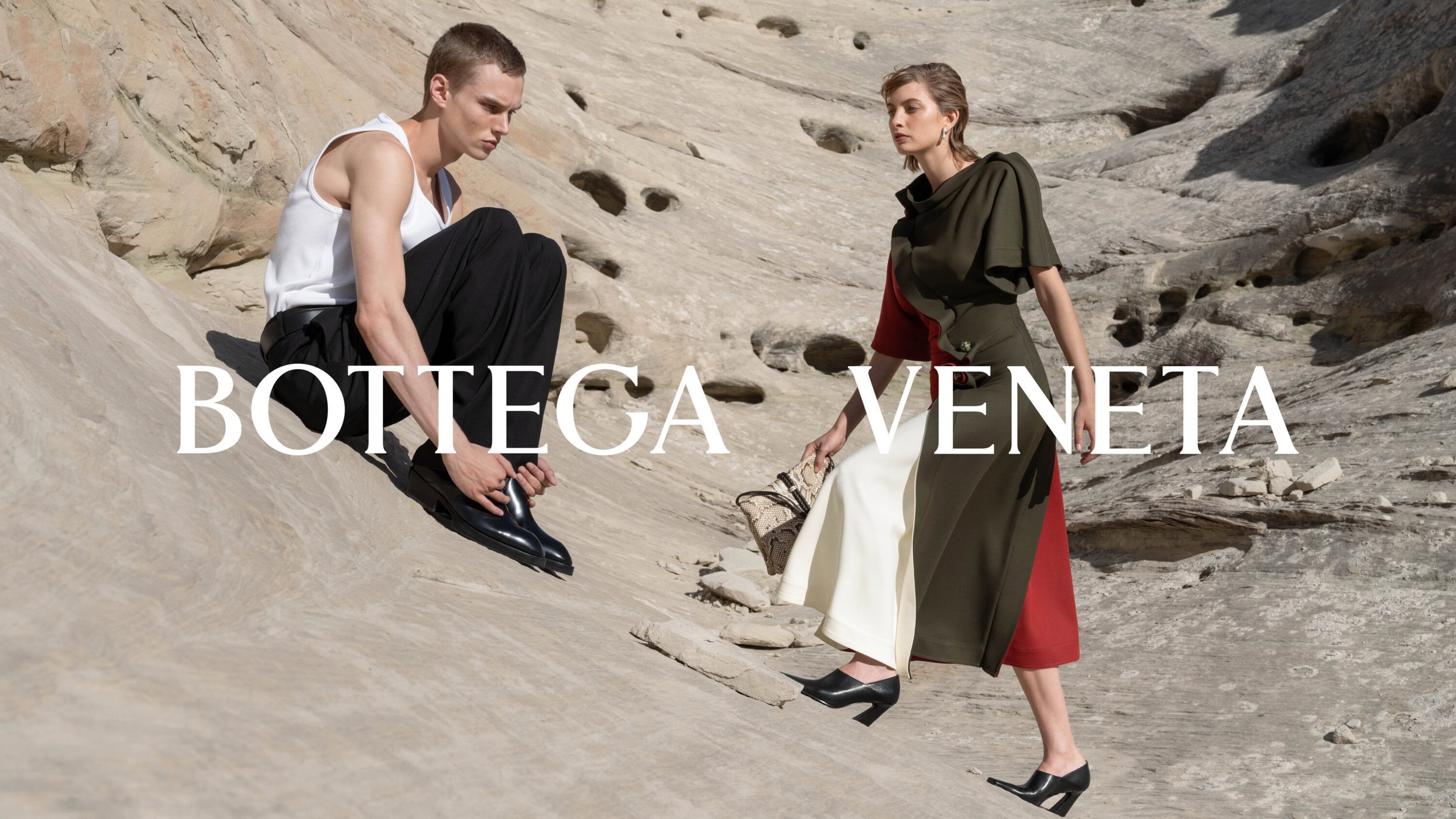 Man in white tank top and woman in structured dress posing on rocky sandstone terrain for Bottega Veneta Winter 2024 campaign