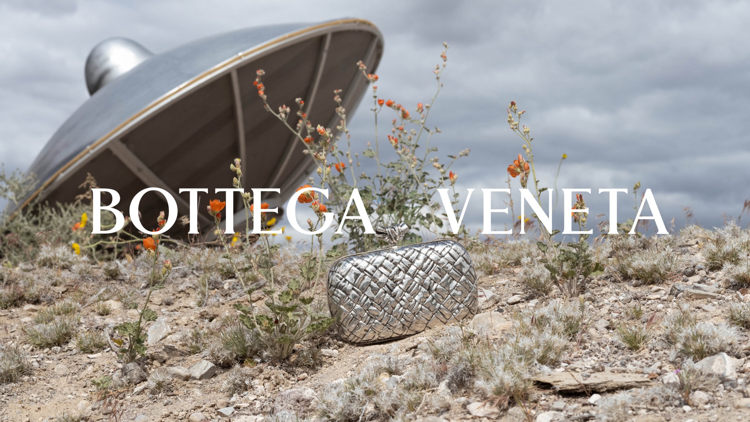Silver woven leather clutch resting on rocky ground with small orange flowers and a large radar dish in the background under an overcast sky.