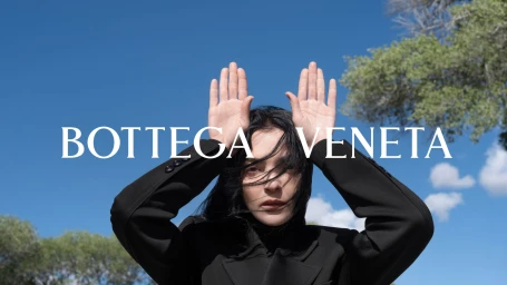 Model in dark jacket holding hands up by head under a bright blue sky for Bottega Veneta
