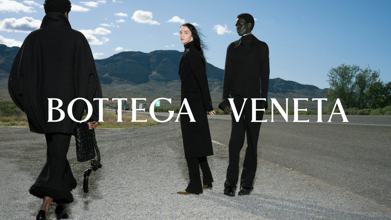 Three models wearing black clothing standing along an asphalt roadside with distant mountains and a cloudy blue sky.