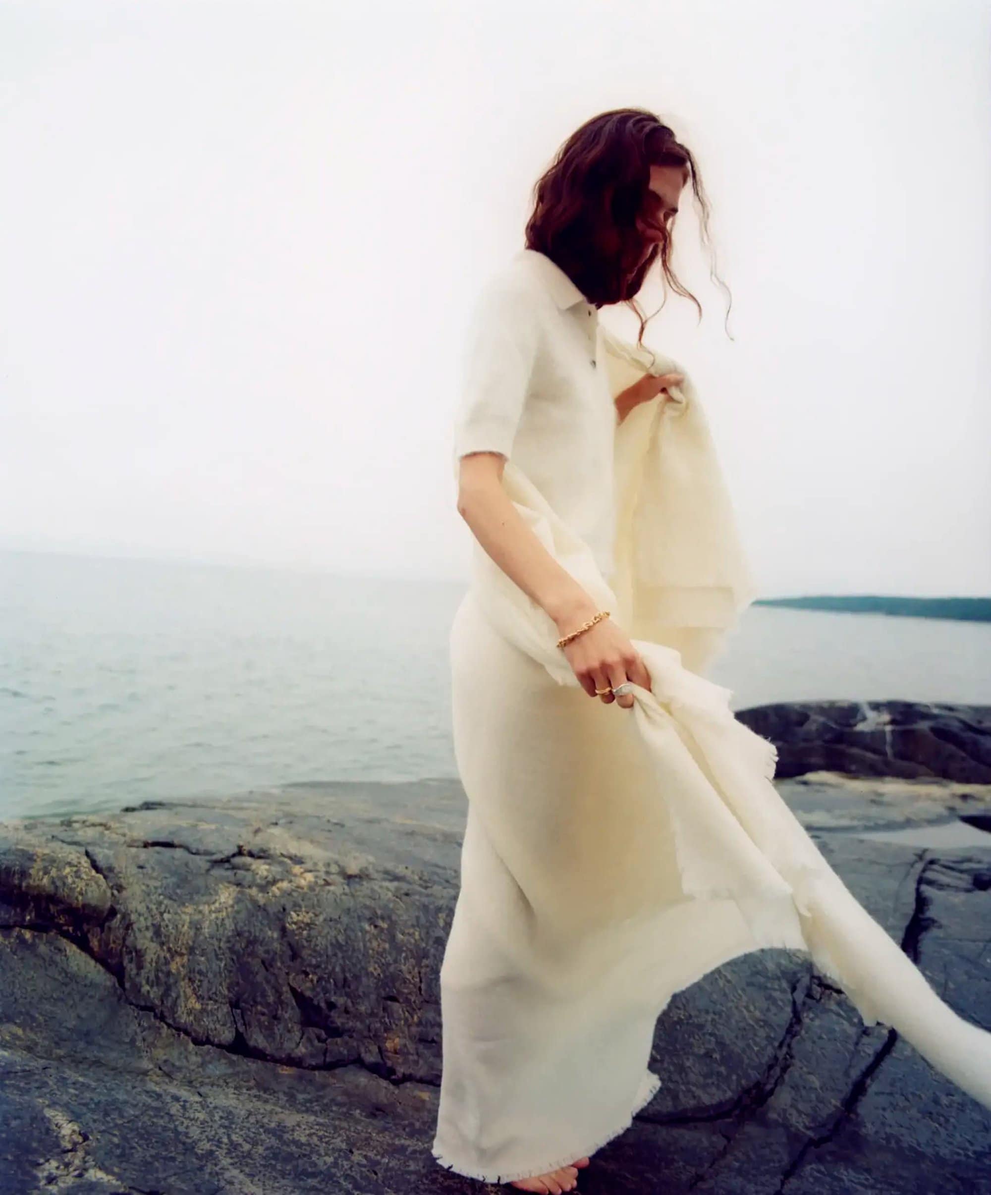 Model in white knit top and skirt standing on dark rocky coast by the ocean