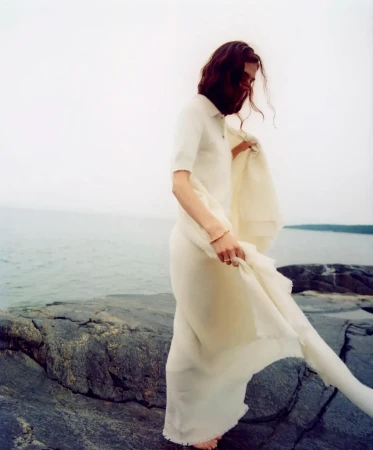 Model in white knit top and skirt standing on dark rocky coast by the ocean