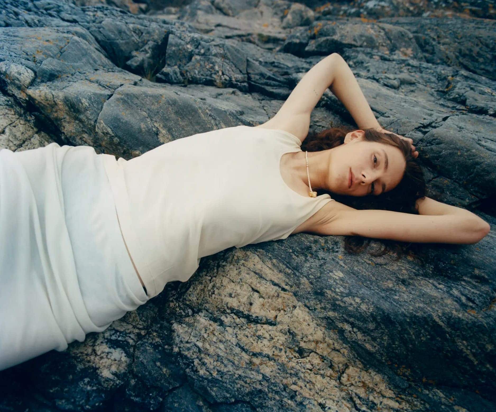 Model lying on dark rocky terrain wearing a light cream tank top and skirt for Simuero Marea Collection