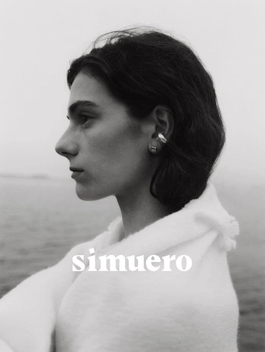 Profile portrait of a woman wearing silver earrings and an off-white textured garment outdoors near water for Simuero