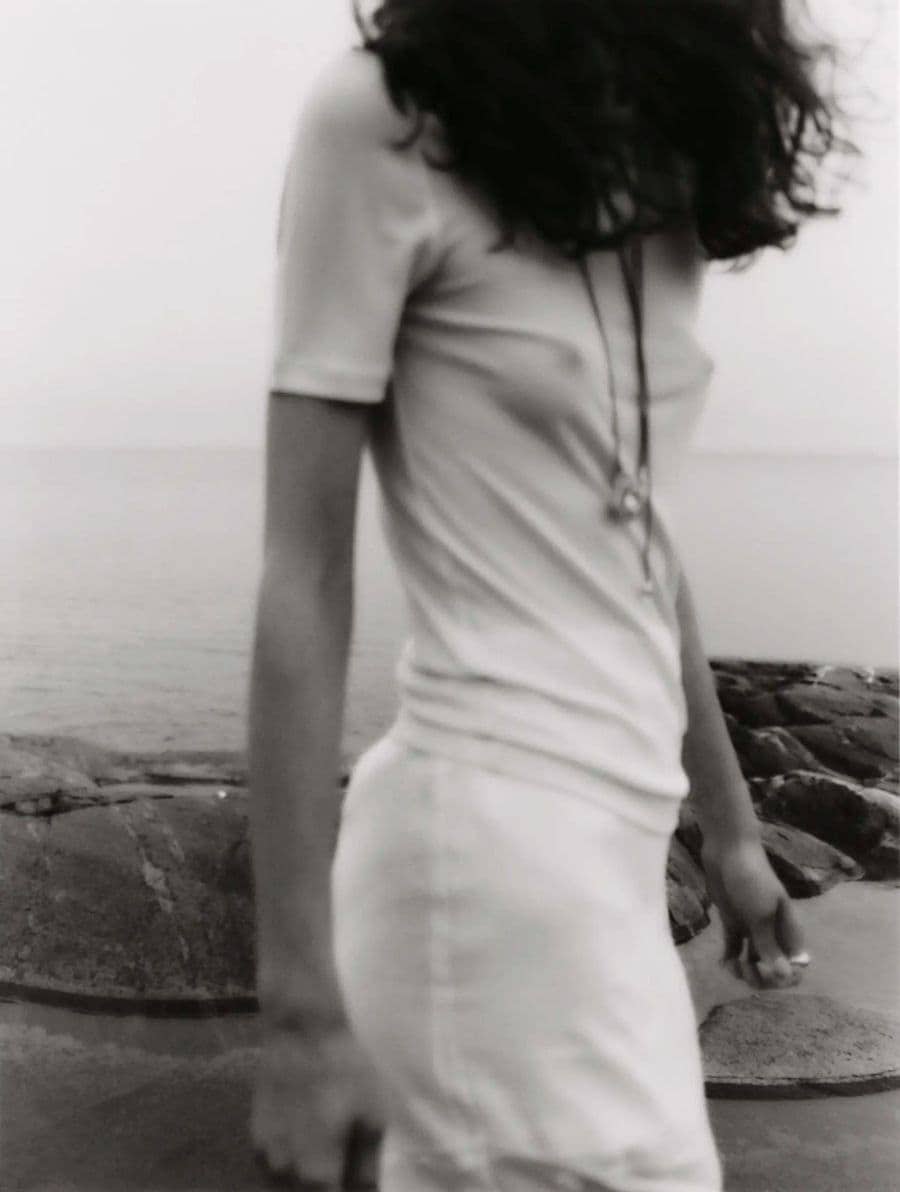 A woman wearing a white t-shirt and white skirt stands on a rocky shore by the water for the Simuero Marea Collection.