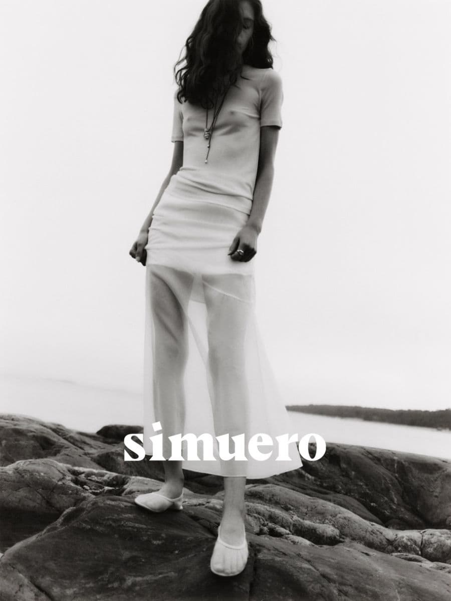 Model standing on dark rocky coastline wearing a light sheer dress and white footwear for Simuero Marea collection